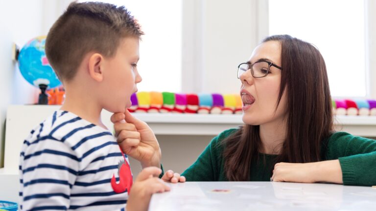 A speech therapist shows a young boy a tongue movement while he watches closely with his hand on his chin.