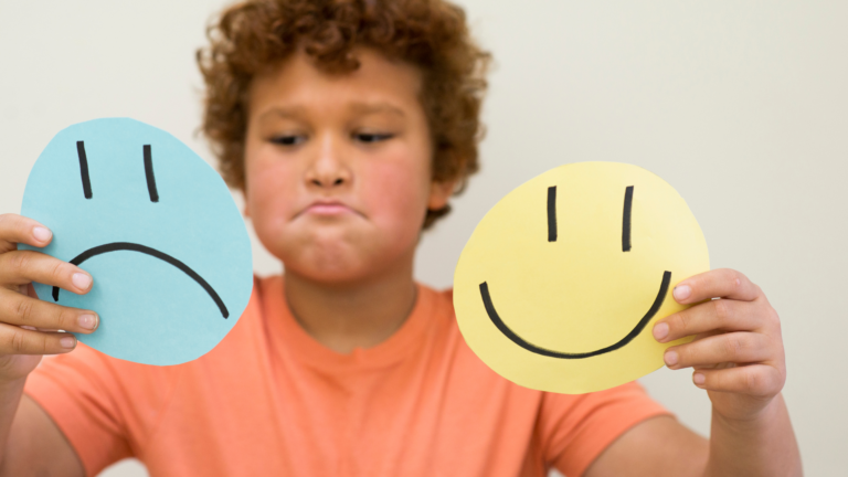 A boy holds up paper cutouts of a sad face and a happy face as he practices naming feelings.